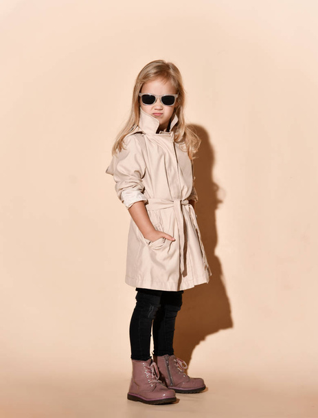 Unhappy blonde kid girl in beige raincoat with rolled up sleeves is standing with her hands in pockets on beige - Photo, image