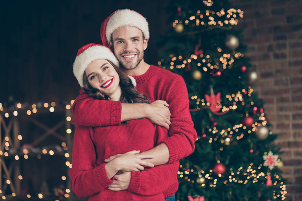 Photo of cute couple spending christmas time eve in decorated garland lights room standing piggyback near x-mas tree indoors wear red pullovers and santa hats - Foto, Imagem