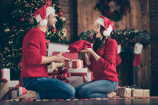 Photo of dreamy couple in decorated garland lights room exchanging holiday gifts sitting cozy on floor near x-mas tree indoors wear red sweaters and santa hats - Foto, afbeelding