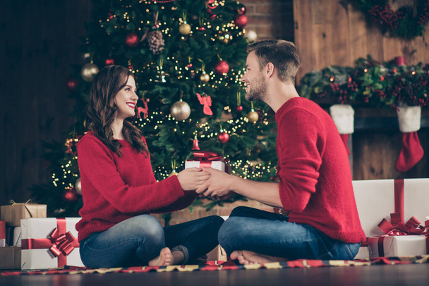Photo of cute couple sharing gifts in x-mas eve spending time in decorated garland lights room sitting cosy on floor near evergreen tree indoors wearing red pullovers - Zdjęcie, obraz