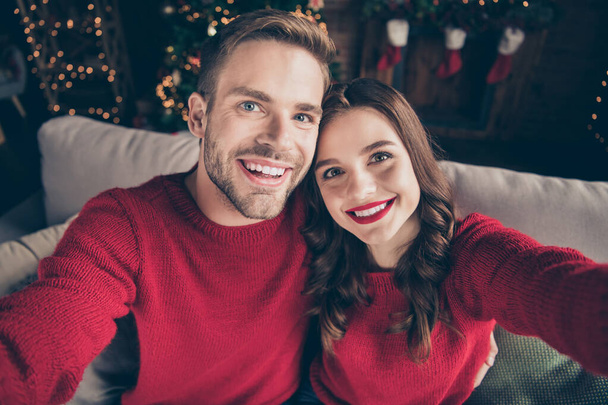 Zdjęcie uroczej pary spędzającej Boże Narodzenie razem robiąc selfie w urządzonym pokoju z lampkami do pończoch w pobliżu nowej choinki w pomieszczeniach noszą czerwone pulowery - Zdjęcie, obraz