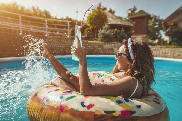 schöne junge Frau im Schwimmbad schwimmt auf aufblasbarem Ring Donut und hat Spaß mit einem Glas Cocktail im Urlaub. - Foto, Bild