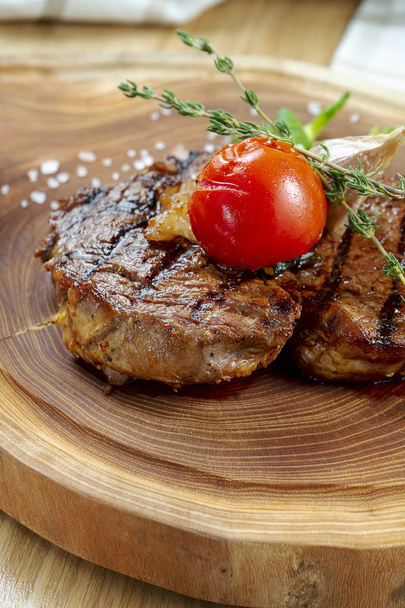 Close up view on tasty grilled steak on wooden board. Composition with cooked beef, herbs and vegetables. Food photo background. Selective focus - Photo, Image