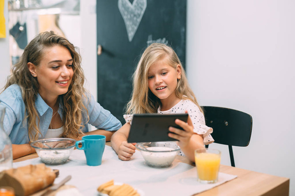 Family time at home. Mother and daughter using tablet while having breakfast. - Foto, afbeelding