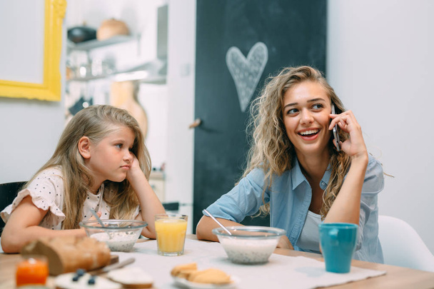 Mother and daughter having breakfast at home, girl boring while woman using smartphone. - Foto, Bild