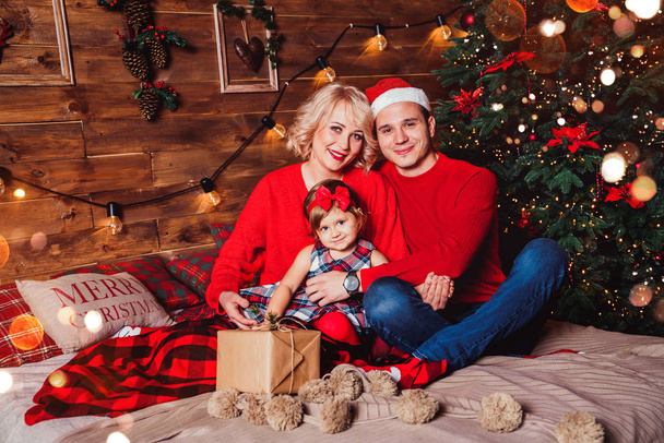 Різдвяна сім'я з дитиною посміхається біля ялинки. Вітальня, прикрашена ялинкою та подарунковими коробками, надає світлу затишну атмосферу. Новорічні свята
 - Фото, зображення