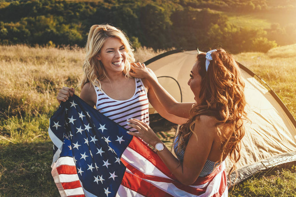 Happy young girl friends enjoys a sunny day in nature. They're holding an american flag in front a campsite tent.  - Photo, image
