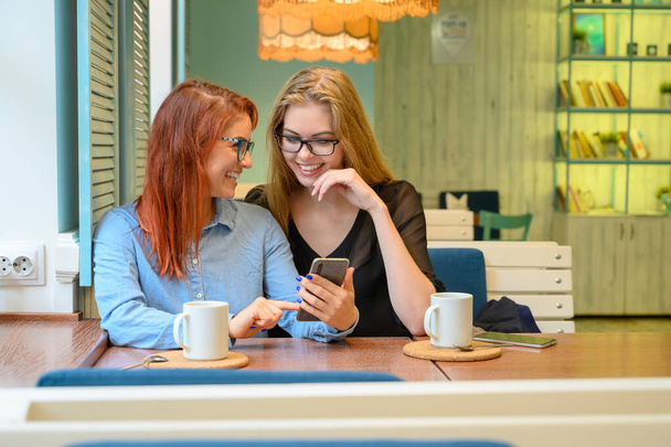Two excited young girls using mobile phones, sitting in a cafe and pointing finger. Red-haired woman in glasses shows her friend funny photos on a smartphone girlfriend blonde. They drink coffee. - Foto, Imagen