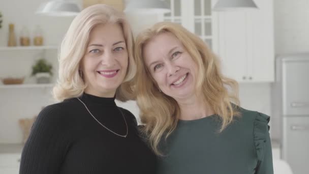 Close-up portrait of two beautiful mature Caucasian women looking at the camera and smiling. Positive attractive ladies standing in luxurious kitchen. - Imágenes, Vídeo