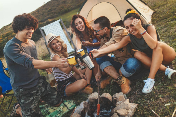 Счастливые молодые друзья наслаждаются солнечным днем в горах. Они смеются и жарят сосиски на палочках у костра возле палатки.
. - Фото, изображение