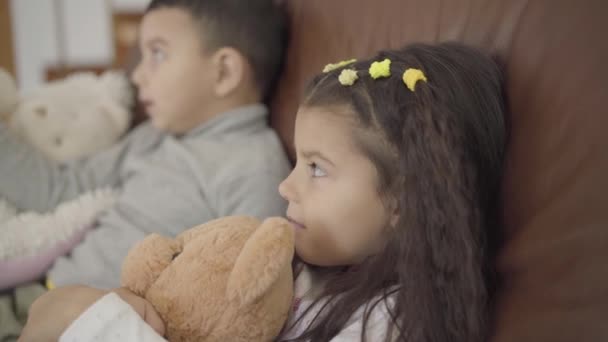 Close-up face of a cute Middle Eastern girl sitting with her brother at the coach and holding teddy bear. Little smiling lady spending evening at cozy home with family. - Video