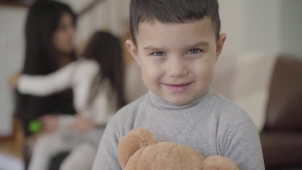 Close-up face of a charming Middle Eastern grey-eyed boy with dark hair holding the teddy bear and looking at the camera. Child posing at home while his sister and mom playing at the background. - Filmati, video