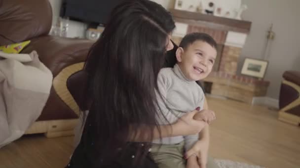 Portrait of a playful Middle Eatern boy imitating airplane flight. Cheerful mother with long dark hair playing with a smiling little boy at home. Camera moving dynamically following people. - Záběry, video