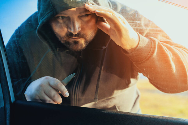 Kriminální osoba v kapuci se dívá dovnitř auta přes sklo nebo okno, zloděj nebo koncepce krádeže auta - Fotografie, Obrázek