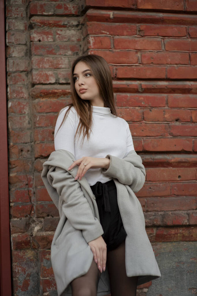 young girl in a gray coat stands on the background of a city street. Girl in a gray coat on a gray background. - Foto, Imagen