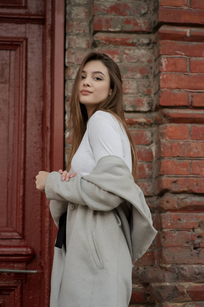 young girl in a gray coat stands on the background of a city street. Girl in a gray coat on a gray background. - Photo, Image