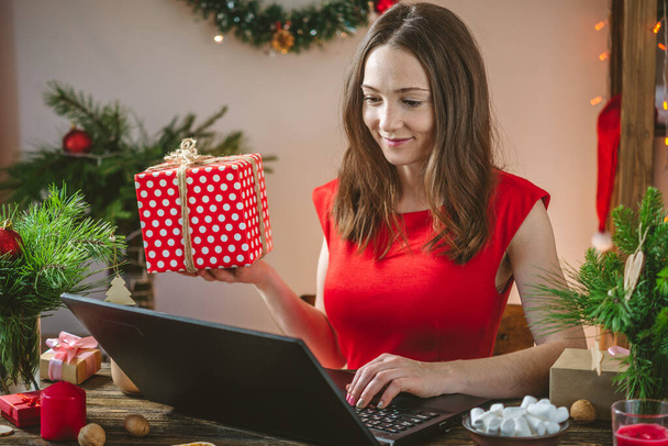 Woman in a red dress is holding a gift box and ordering purchases on laptop. Online shopping for Christmas holidays. - Fotografie, Obrázek