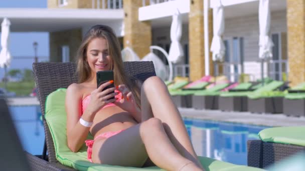 nyári üdülőhely, vidám vonzó lány fürdőruhában mobiltelefonnal a kezében pihentető nyugágyon medencével drága üdülőhelyen nyaralás közben - Felvétel, videó