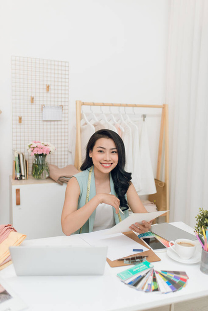Модельер на работе. Счастливая молодая азиатская женщина рисует, сидя на своем рабочем месте в мастерской моды
 - Фото, изображение