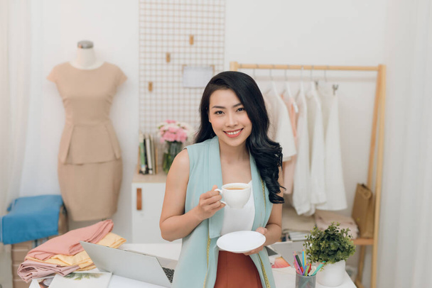 Happy young Asian fashion designer business drinking coffee at her workshop/store - 写真・画像