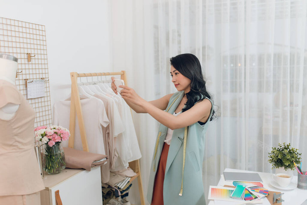Красивая молодая азиатская модельер делает фото работы с помощью смартфона в студии портнихи
. - Фото, изображение