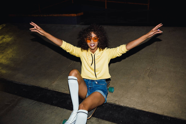 Image of african american girl riding on skateboard at night out - 写真・画像