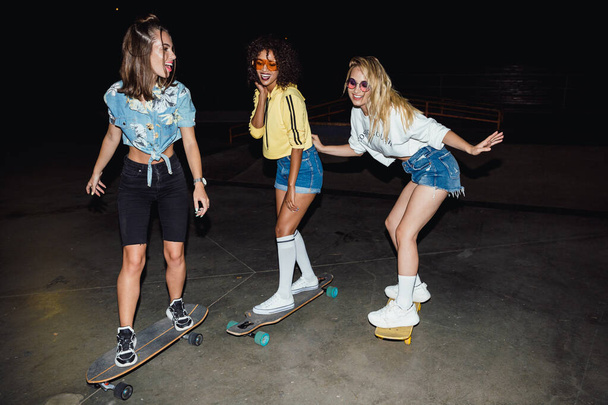 Image of stylish multinational girls riding skateboards at night - Фото, зображення