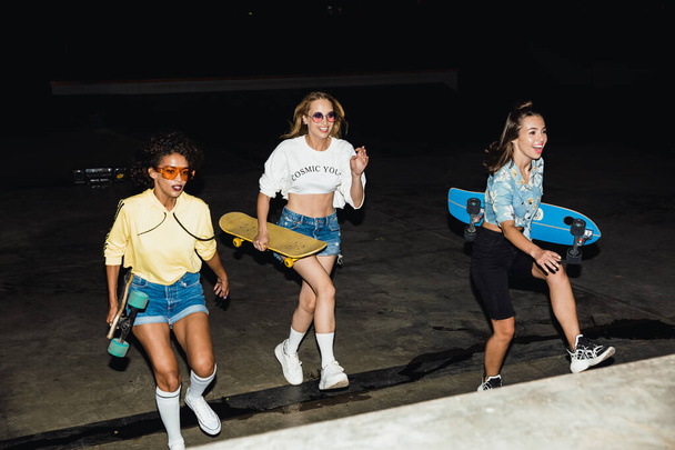 Image of fashion multiethnic girls smiling and riding skateboard - Valokuva, kuva