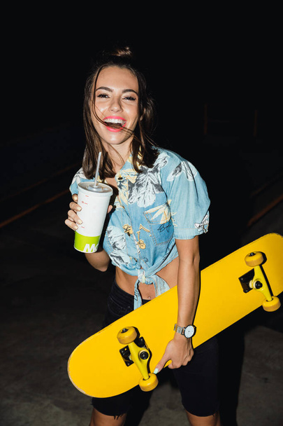 Image of brunette girl drinking soda and holding skateboard at n - Zdjęcie, obraz