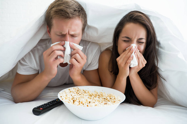 young couple handsome man and beautiful woman watching TV in bed with pop corn looking sick bowing their noses - Фото, зображення