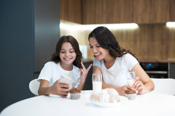 young mother with her teenager daughter watching photoes on the phone while having cupcakes with milk for breakfast both smiling - Photo, Image