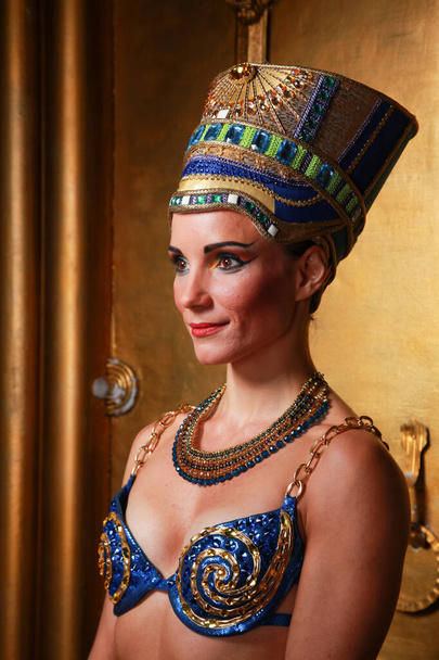Beautiful thin Egyptian woman with pronounced cheekbones in a huge gold headdress without hair in a blue swimsuit on the background in the Studio - Photo, image