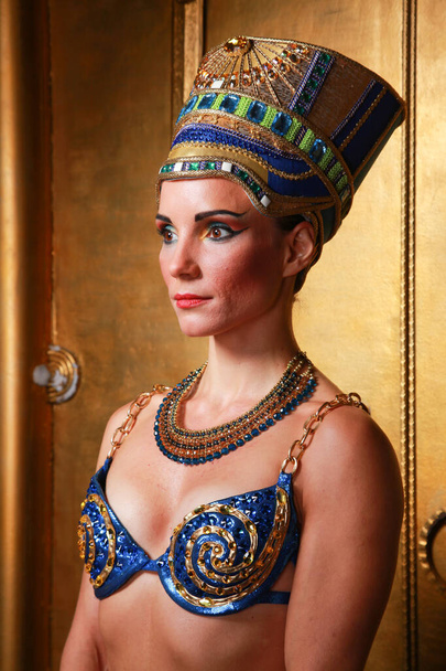 Bella donna egiziana sottile con zigomi pronunciati in un enorme copricapo d'oro senza capelli in un costume da bagno blu sullo sfondo in Studio
 - Foto, immagini