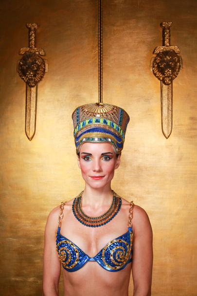 schöne dünne Ägypterin mit ausgeprägten Wangenknochen in einem riesigen goldenen Kopfschmuck ohne Haare in einem blauen Badeanzug auf dem Hintergrund im Studio - Foto, Bild
