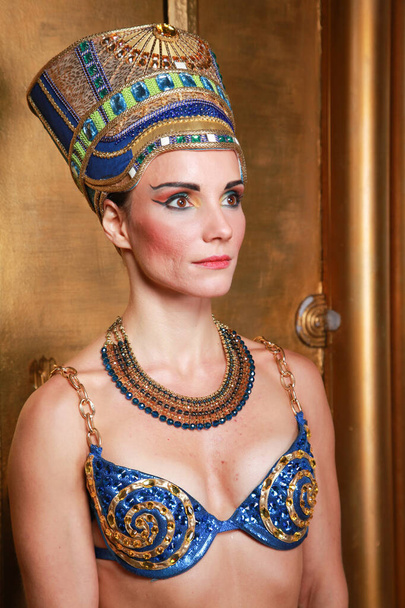 Beautiful thin Egyptian woman with pronounced cheekbones in a huge gold headdress without hair in a blue swimsuit on the background in the Studio - Fotografie, Obrázek