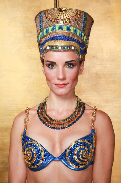 Mulher egípcia fina bonita com maçãs do rosto pronunciadas em uma enorme cobertura para a cabeça de ouro sem cabelo em um maiô azul no fundo no estúdio
 - Foto, Imagem