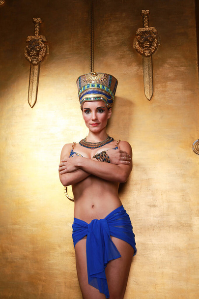 Beautiful thin Egyptian woman with pronounced cheekbones in a huge gold headdress without hair in a blue swimsuit on the background in the Studio - Photo, Image