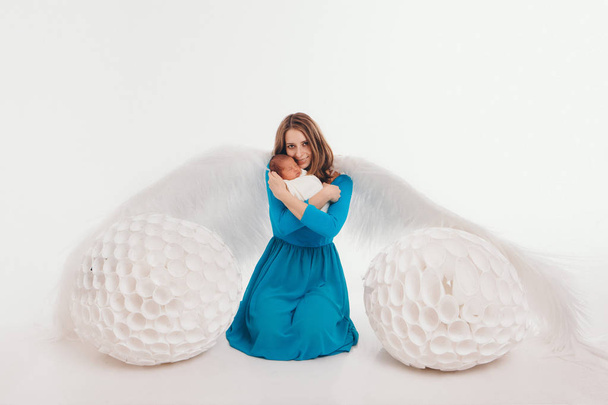 A girl in a blue dress with angel wings holding a newborn baby on a white background. An angel-woman and child have big white balls. The concept of Christmas, mystical creatures, Halloween - Photo, Image