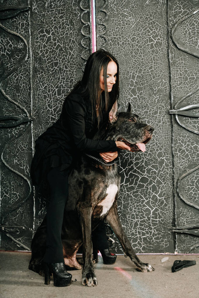 schöne junge Frau posiert mit ihrem großen Hund im Studio in der Nähe großer schwarzer Tore Hintergrund - Foto, Bild
