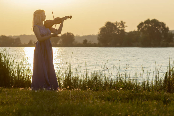 romantic silhouette of young woman with a violin at dawn on river bank, elegant girl playing a musical instrument on nature, concept music and inspiration - Фото, зображення