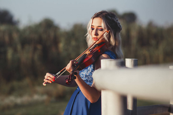 portrait of a beautiful girl in a long dress with a violin near a wooden painted fence, a young woman walks with a musical instrument outdoors - Foto, Bild