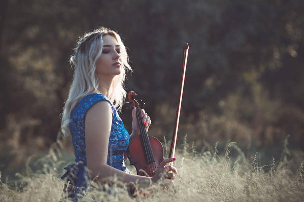 portrait of a young fabulously beautiful girl in a dress with a violin sitting in dry grass on meadoe at the dawn, woman playing a musical instrument with inspiration relaxing on nature - Foto, immagini
