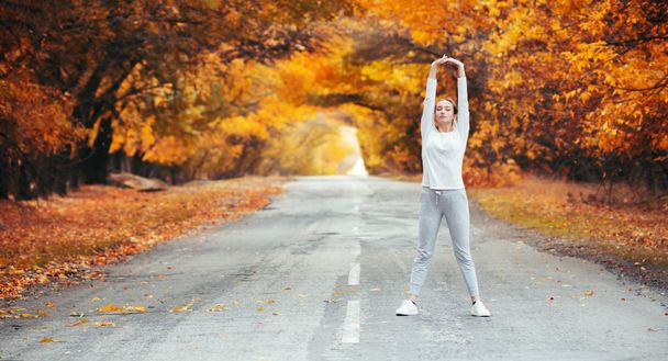 slim young woman in sportswear exercises and stretches hands up standing on asphalt road, girl engaged in sport outdoors on background of autumn foliage, concept healthy lifestyle and female beauty - Photo, Image