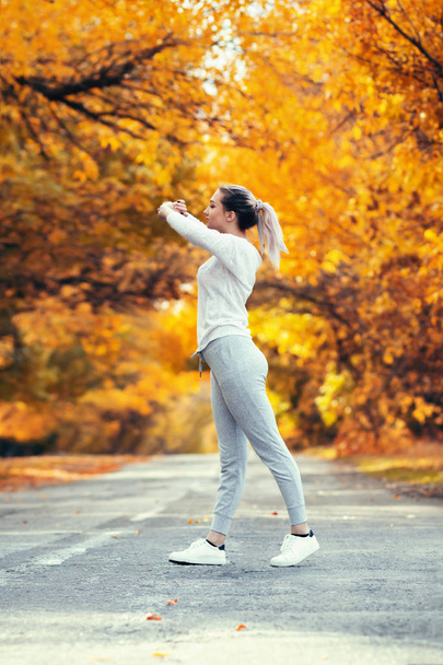 slim young woman in sportswear exercises and stretches hands up satanding on asphalt road, girl engaged in sport outdoors on background of autumn foliage, concept healthy lifestyle and female beauty - Photo, Image