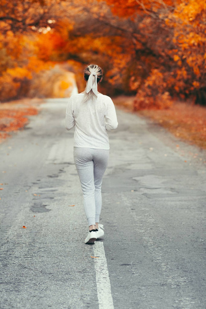 back of young woman in sportswear jogging on asphalt road on background of red foliage, girl engaged in sport outdoors on autumn day, concept healthy lifestyle and hobby - Photo, Image