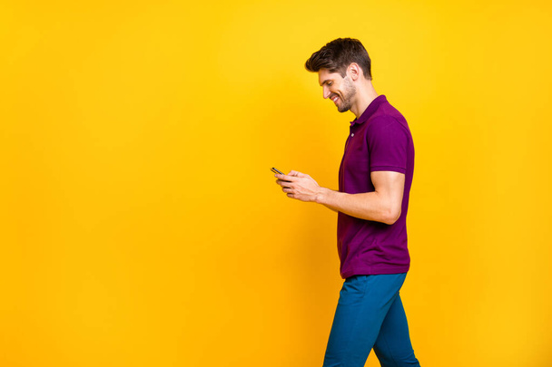 Photo of cheerful positive trendy white man browsing through his telephone talking with friends wearing blue pants trousers going moving side profile isolated over vivid color background - Zdjęcie, obraz