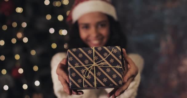 Close Up of Pretty Smiling Mulatto Girl in Santas Hat Gives and Takes Back the Christmas Present Box to Camera, Standing at Decorative Lighting at the Background. Winter Holiday Concept. - Footage, Video