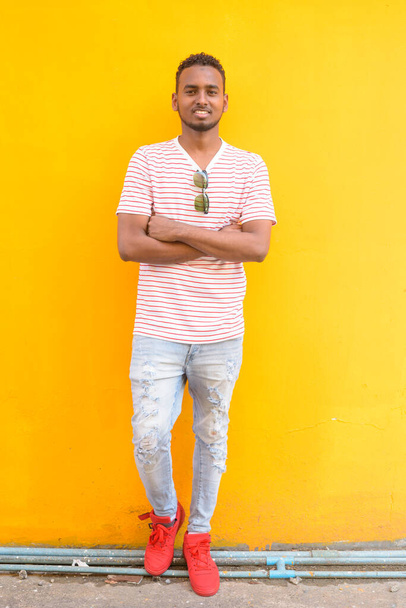 Koko kehon laukaus onnellinen nuori afrikkalainen mies hymyilee kädet ristissä keltaista seinää vasten
 - Valokuva, kuva