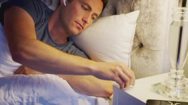Man in bed putting in wireless earphones connected to mobile phone before going to sleep - shot in slow motion - Imágenes, Vídeo