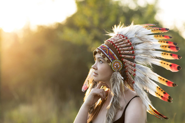 Fiatal gyönyörű lány őshonos amerikai fejdísszel a természetben naplementekor. Amerikai indiai háttér. - Fotó, kép